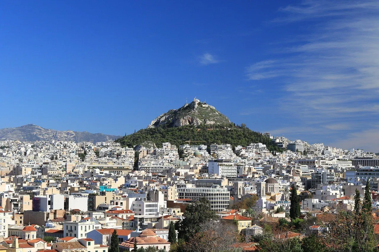 Monte Licabeto com capela branca no cume, vegetação na encosta e prédios de Atenas à frente.
