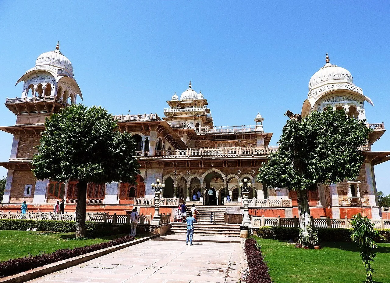 Exterior do Albert Hall em Jaipur, com cúpulas claras, árvores, gramado e um caminho até a escadaria.