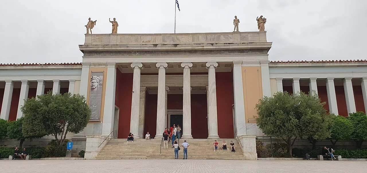 Fachada do Museu Arqueológico Nacional de Atenas, com colunas, escadaria e árvores laterais sob céu nublado.