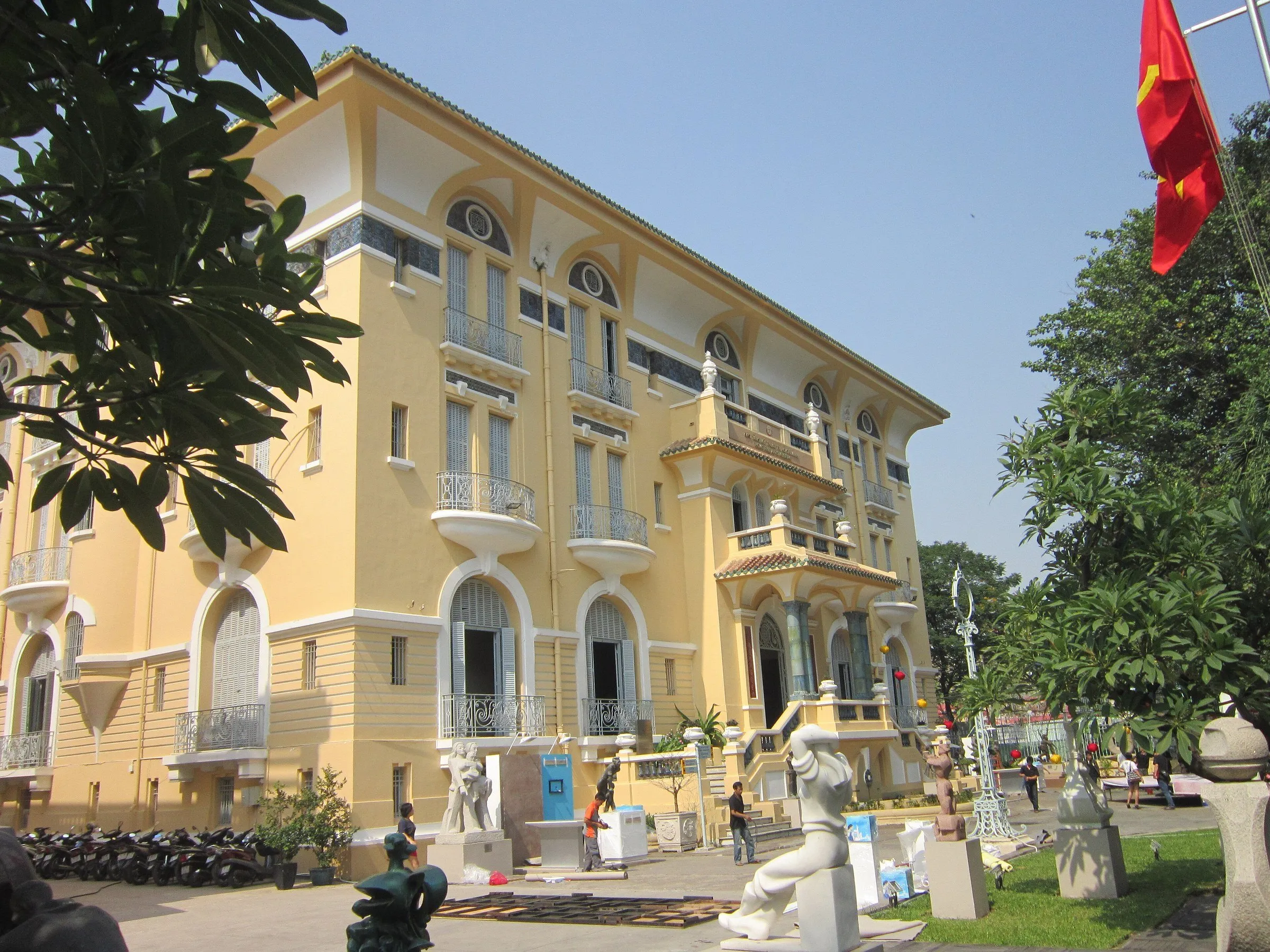 Edifício amarelo do Museu de Belas Artes de Ho Chi Minh com janelas em arco, varandas e esculturas no pátio.
