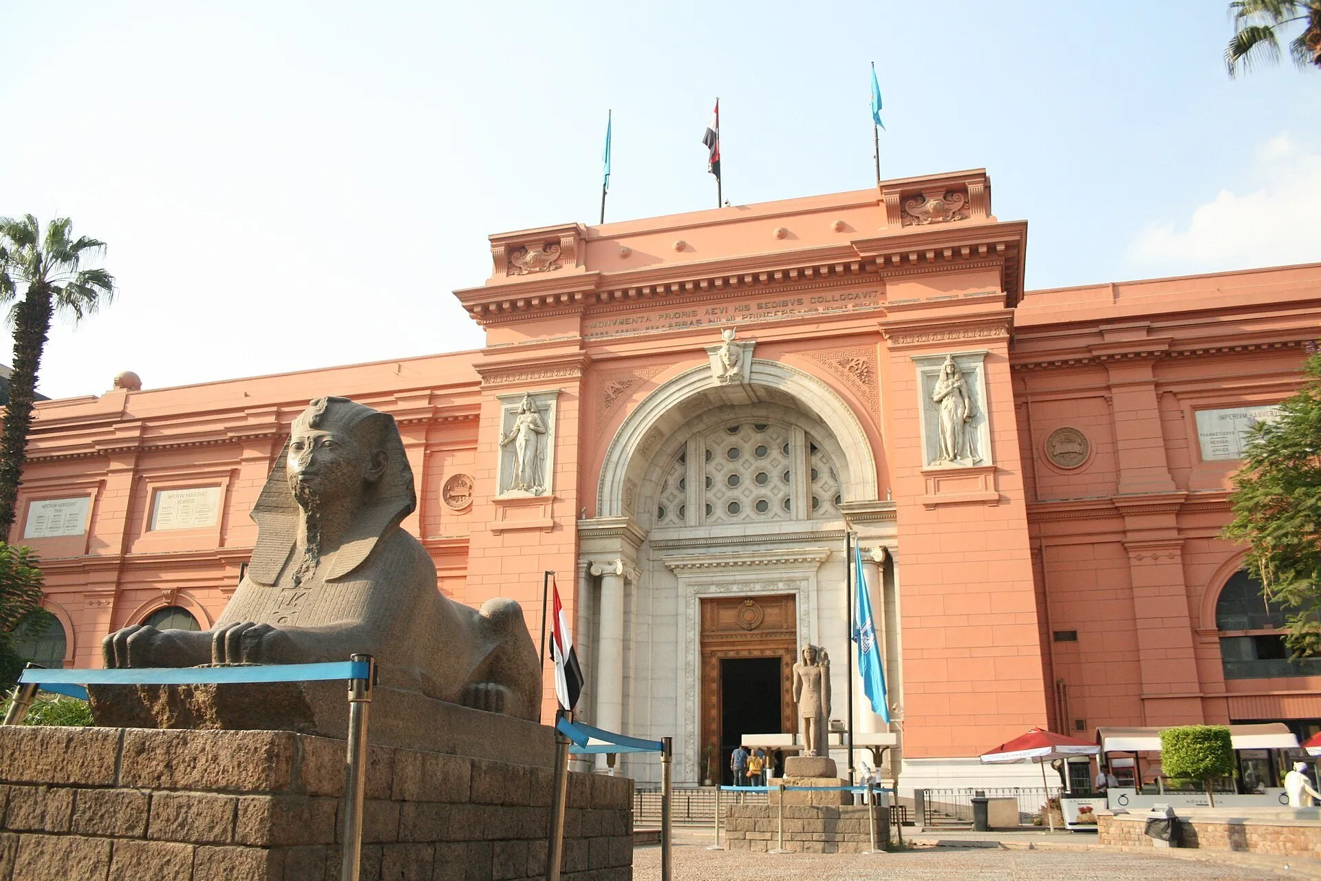 Fachada rosada do Museu Egípcio de Tahrir, com portal central, bandeiras e esfinge de pedra à frente.