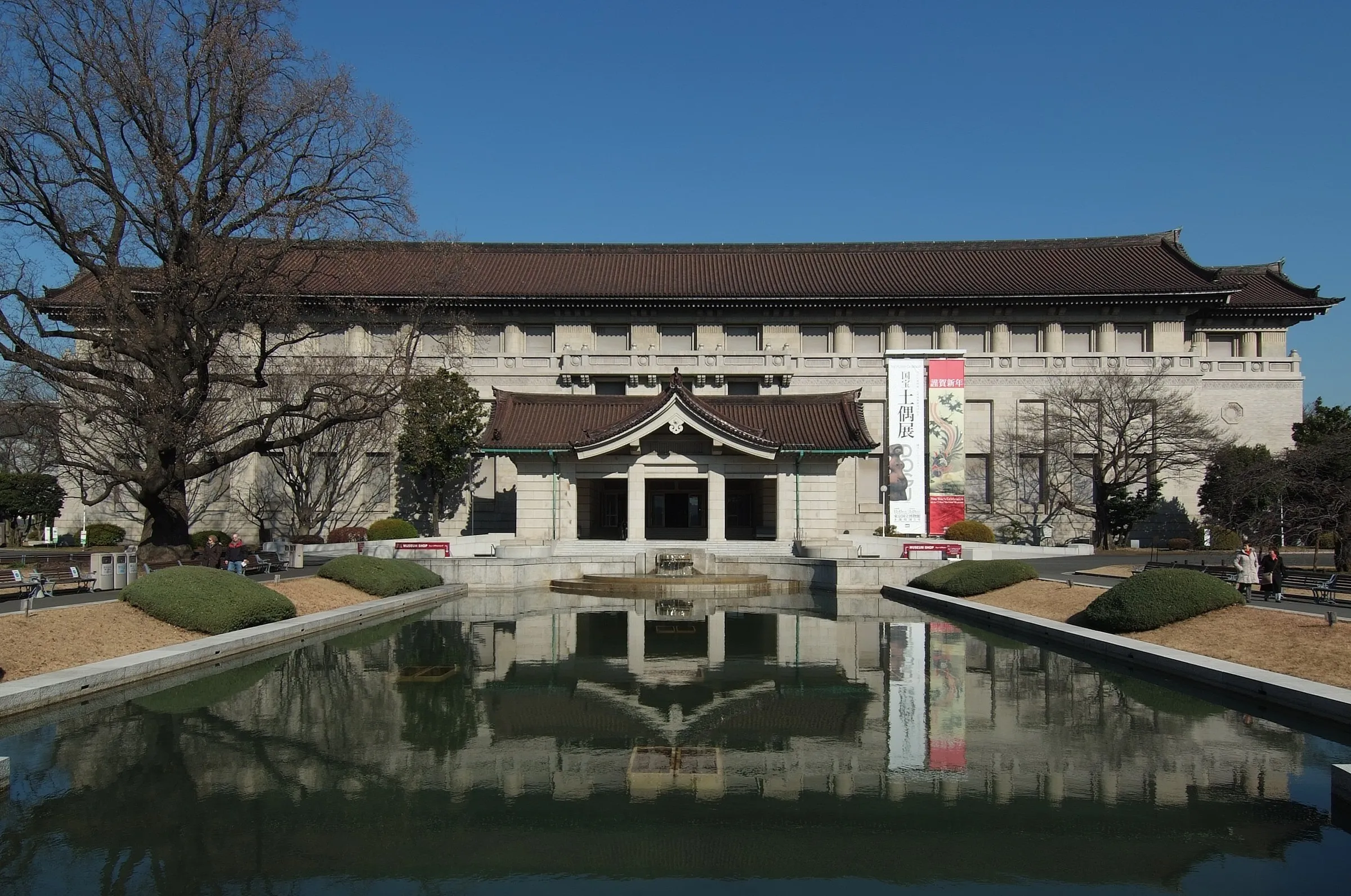 Fachada do Honkan do Museu Nacional de Tóquio refletida no espelho d’água, sob céu azul.