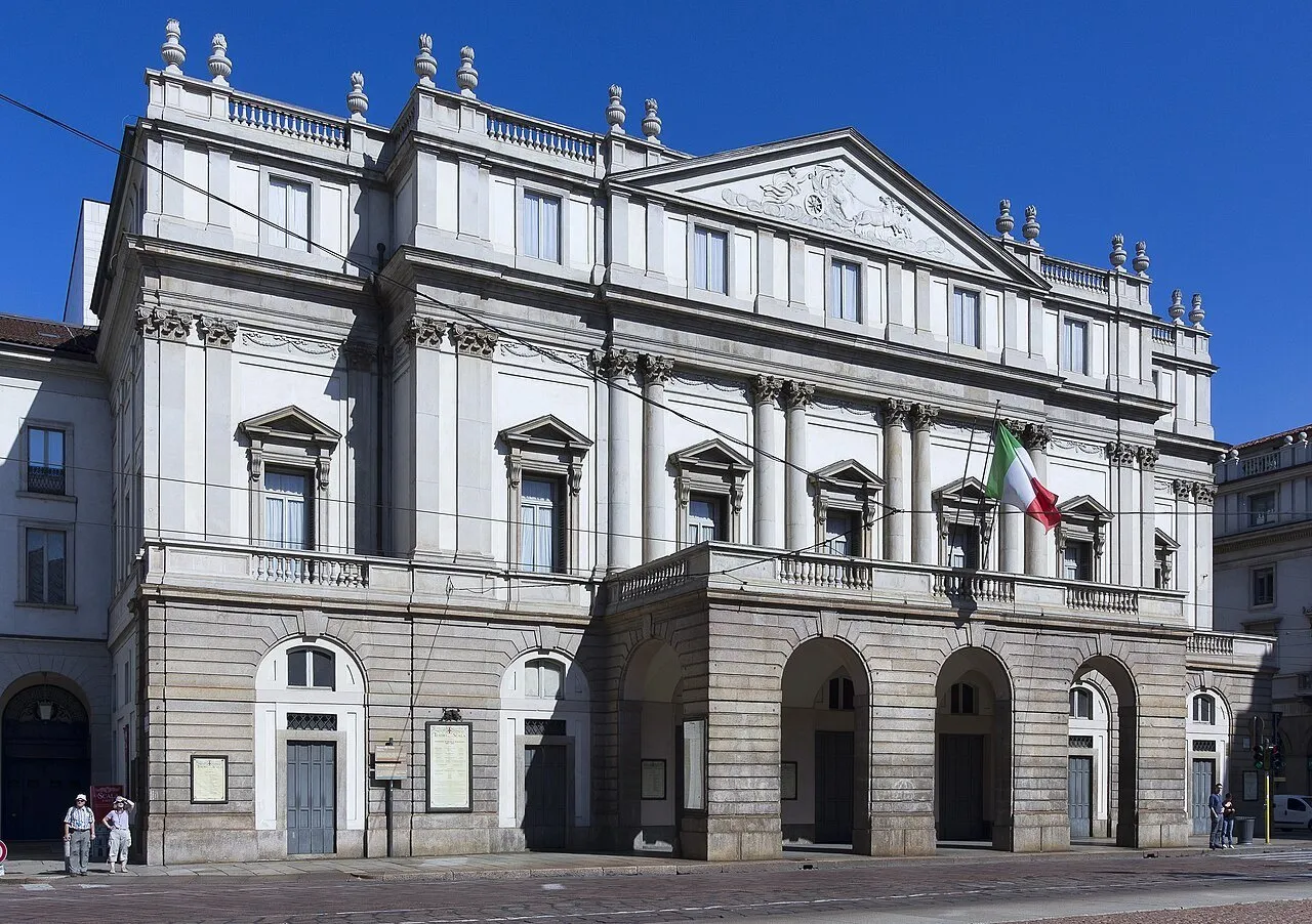 Fachada clara do Teatro alla Scala com pórtico, bandeira italiana e poucos pedestres sob céu azul.