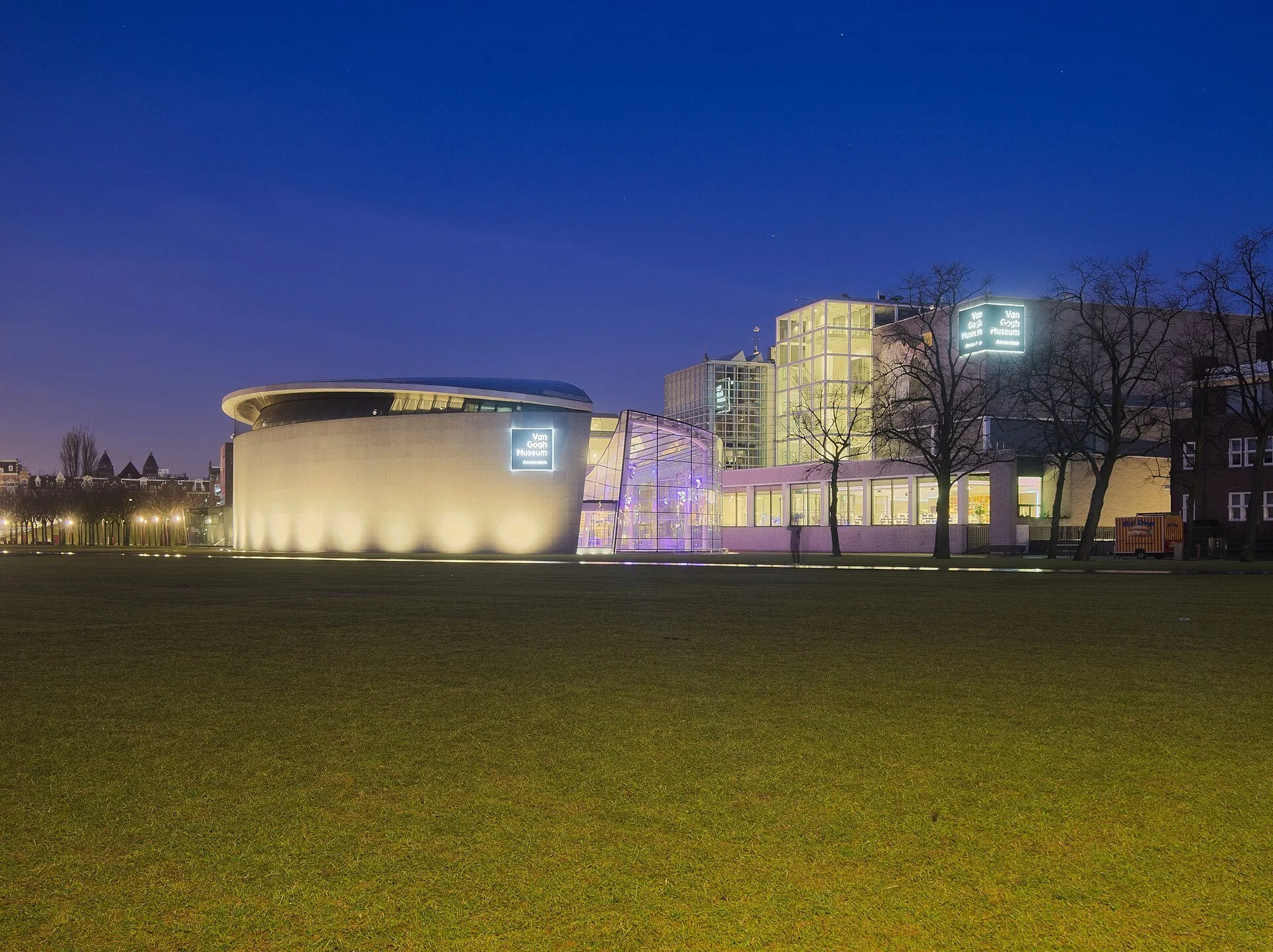 Museu Van Gogh iluminado à noite, com volume curvo, entrada envidraçada e gramado à frente.