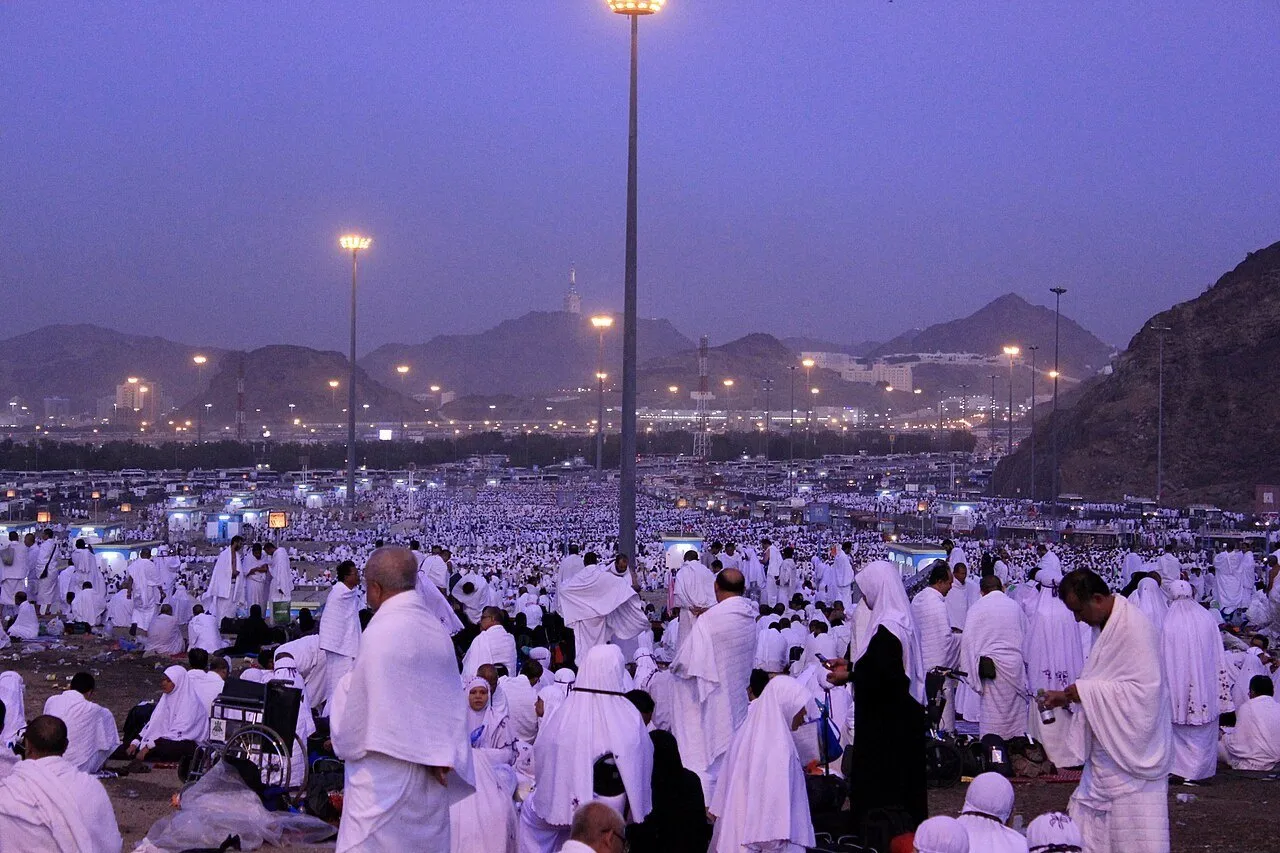 Vista ampla de Muzdalifah na luz azul do amanhecer, com peregrinos na planície, postes e montanhas ao fundo.