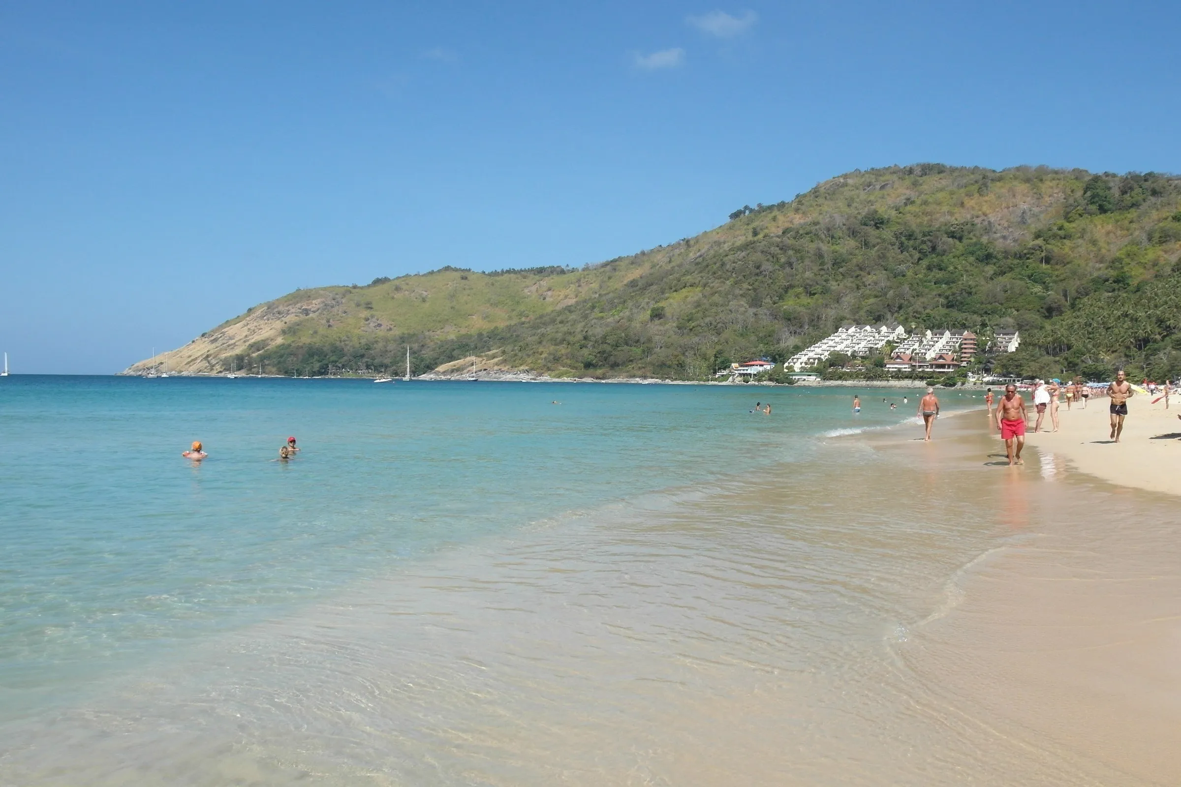Praia de Nai Harn, em Phuket, com areia clara e água turquesa à esquerda, banhistas ao longe e morro verde ao fundo.