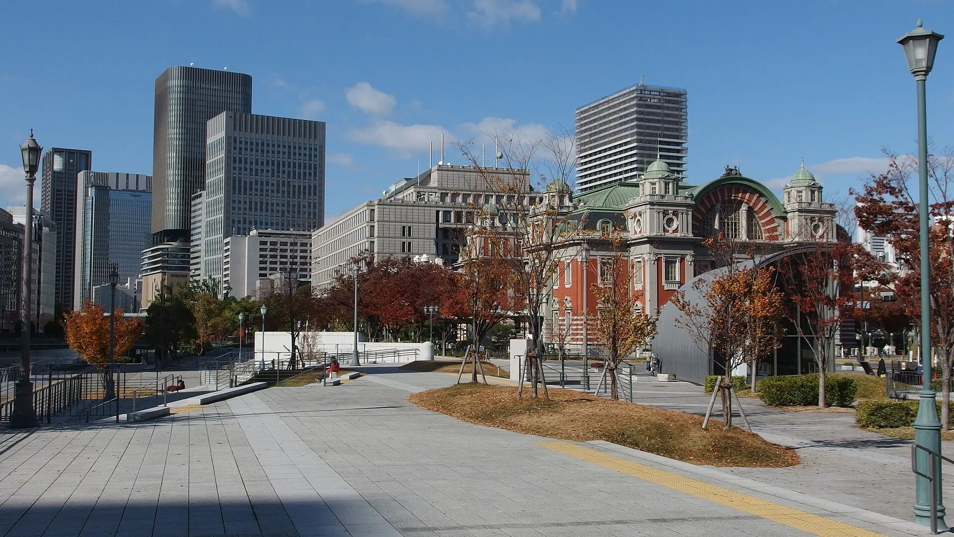 Parque de Nakanoshima com caminhos pavimentados, árvores de outono e o salão público de tijolos ao fundo.