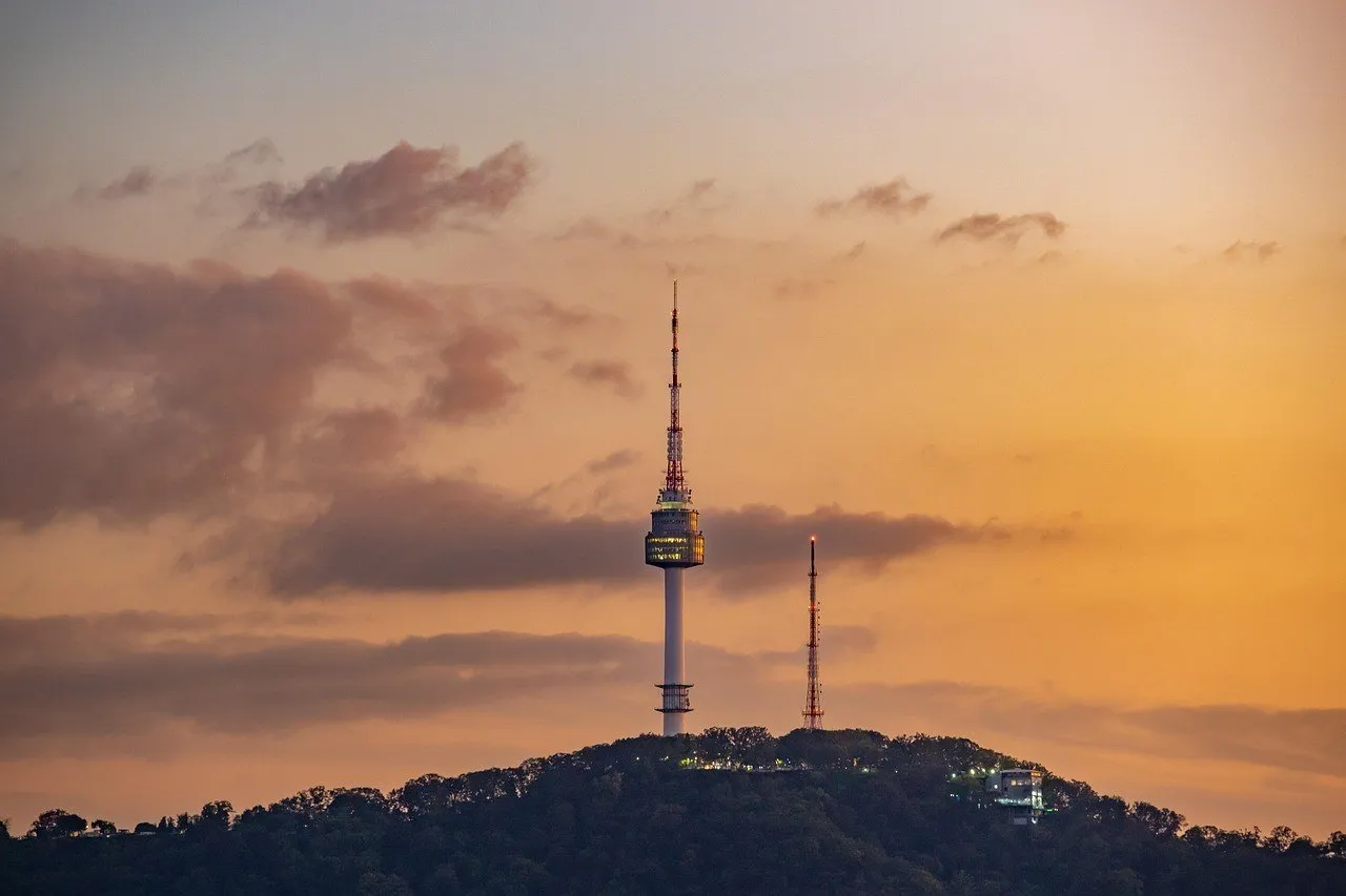 N Seoul Tower no morro arborizado de Namsan, sob céu alaranjado com nuvens.