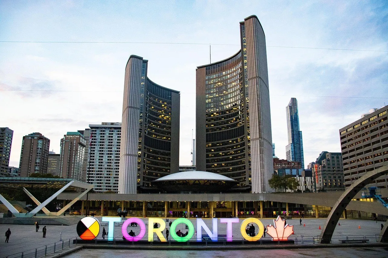 Letreiro TORONTO iluminado em várias cores diante das duas torres curvas da prefeitura ao entardecer.