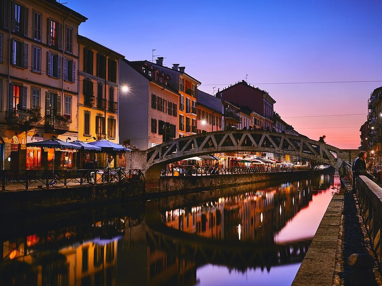 Ponte Alda Merini sobre o Naviglio Grande, com fachadas iluminadas refletidas na água ao entardecer.