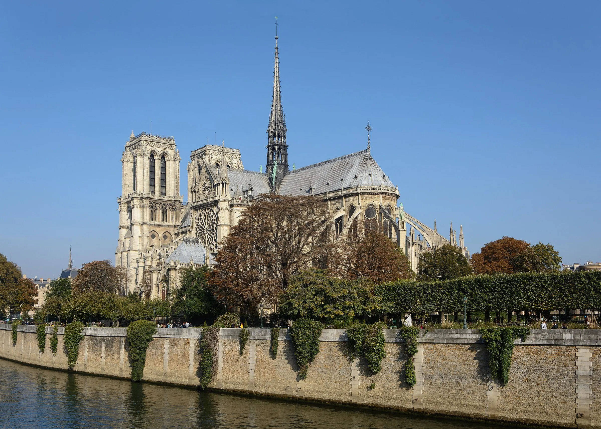Catedral de Notre-Dame vista do Sena, com as duas torres, a agulha e as árvores do cais sob céu azul limpo.
