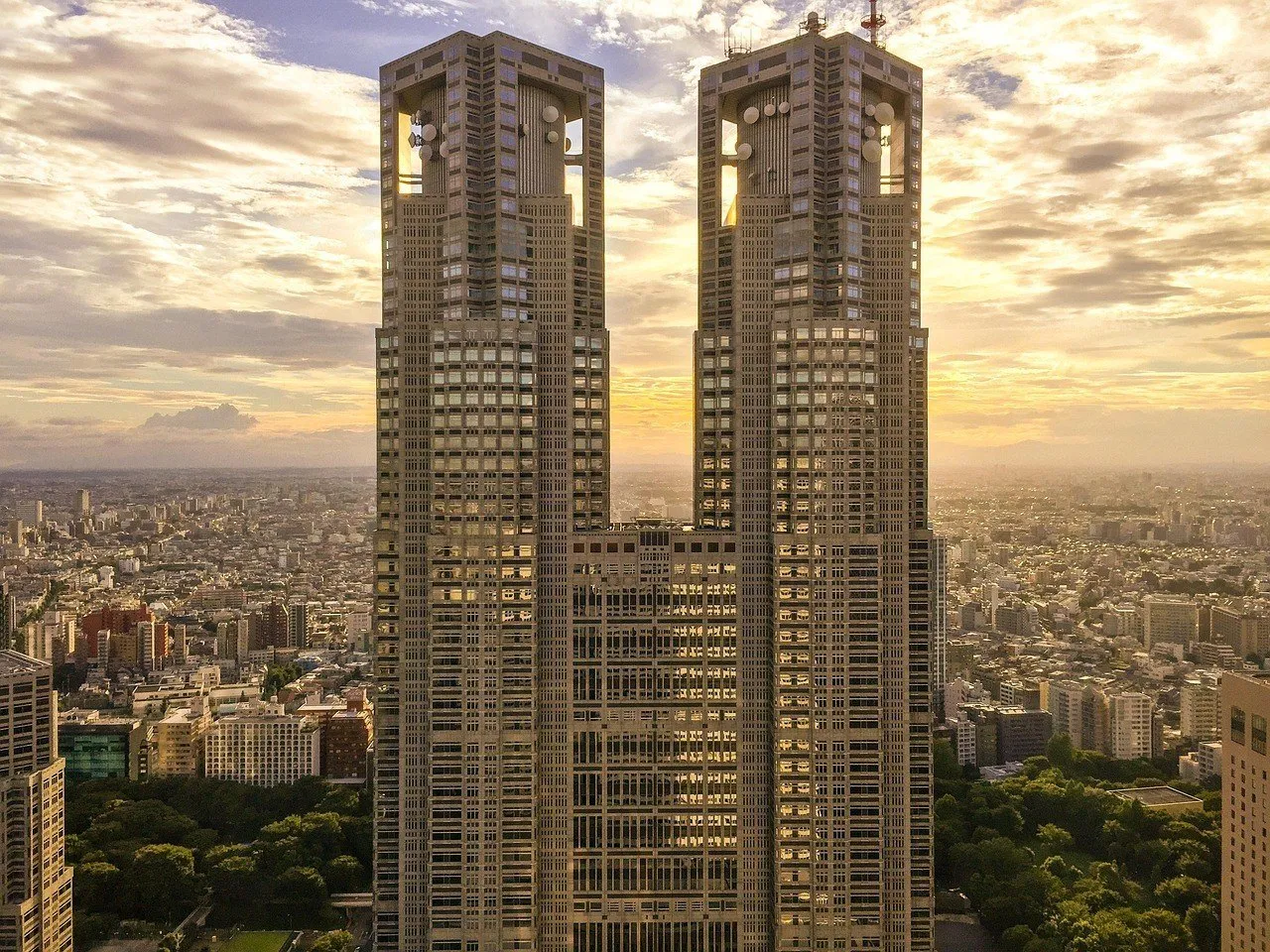 Torres gêmeas do Governo Metropolitano de Tóquio vistas de frente ao entardecer, com prédios ao fundo.