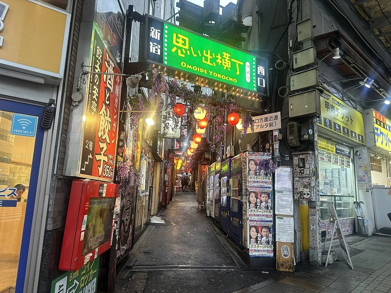 Entrada de Omoide Yokocho com placa verde, lanternas acesas, pequenas fachadas e corredor estreito.