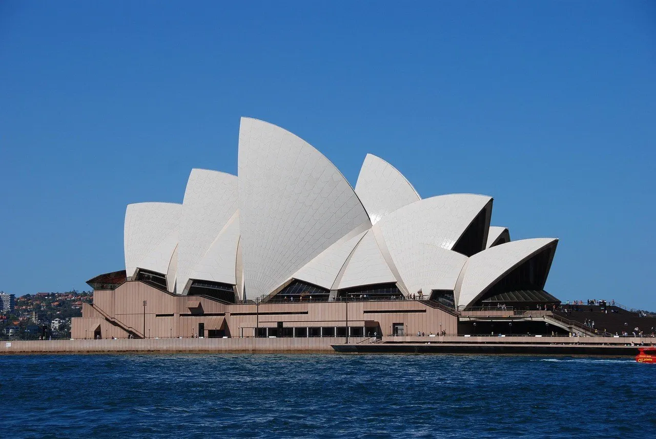 Ópera de Sydney vista da água, com coberturas brancas, base de pedra e céu azul.