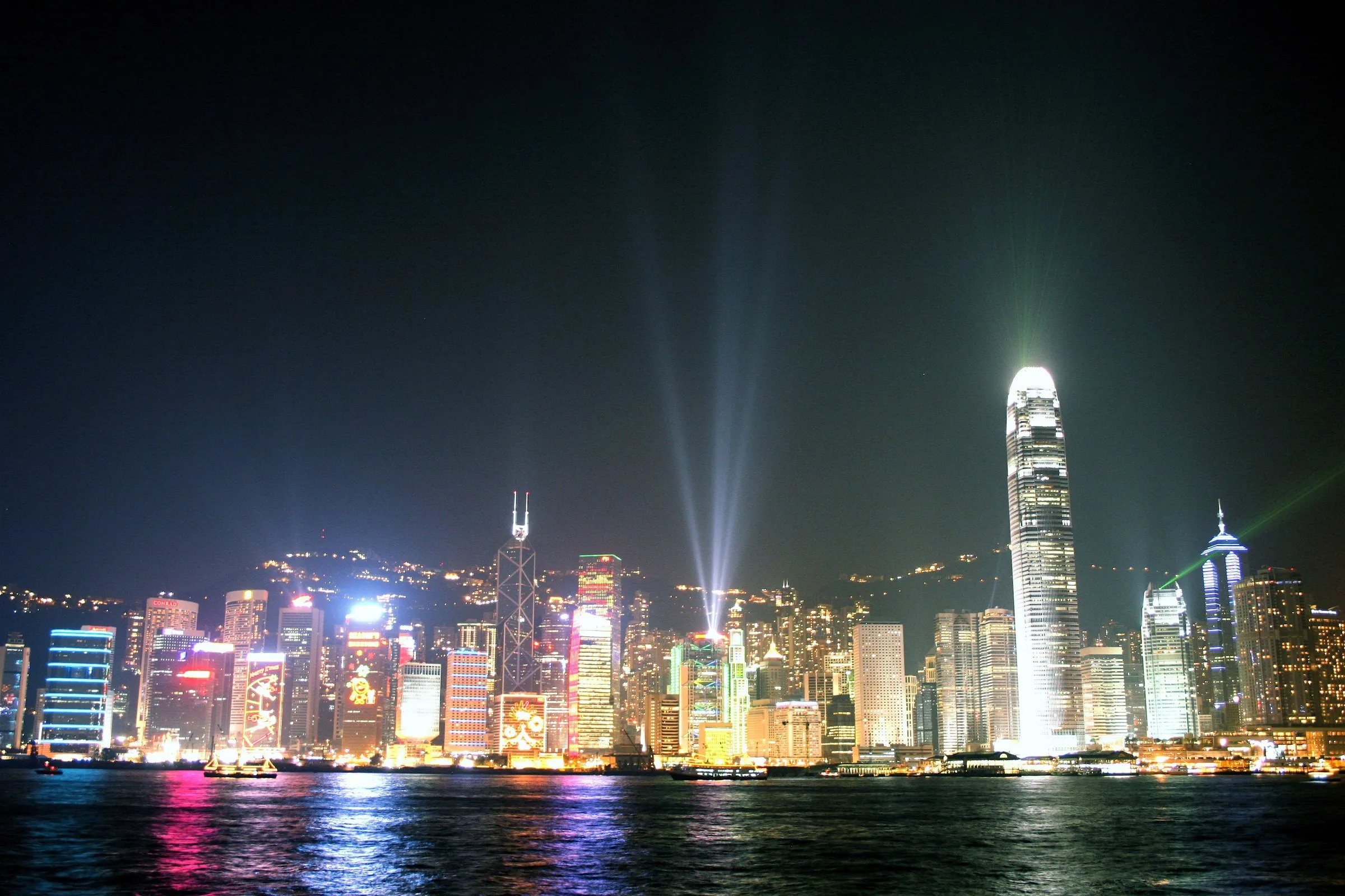 Skyline noturno da ilha de Hong Kong visto do outro lado do porto, com feixes de laser sobre os prédios e reflexos na água.