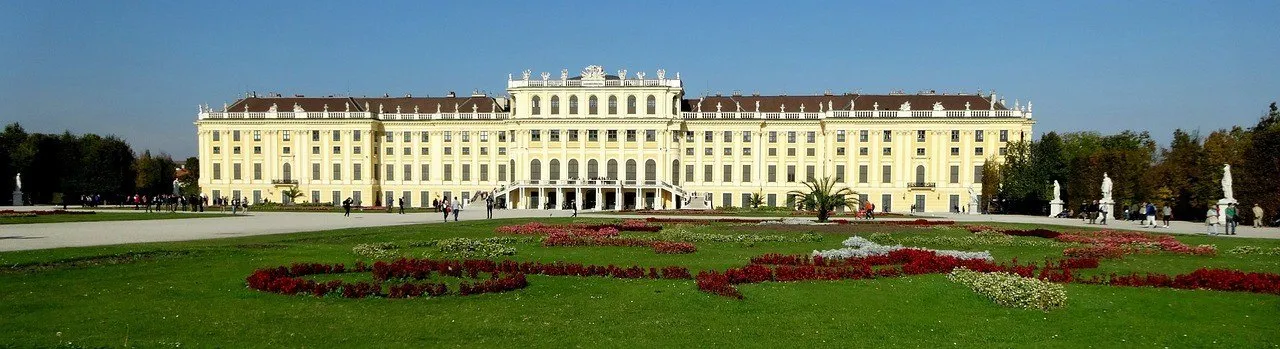 Fachada amarela do Palácio de Schönbrunn diante de gramado e canteiros floridos, sob céu azul.