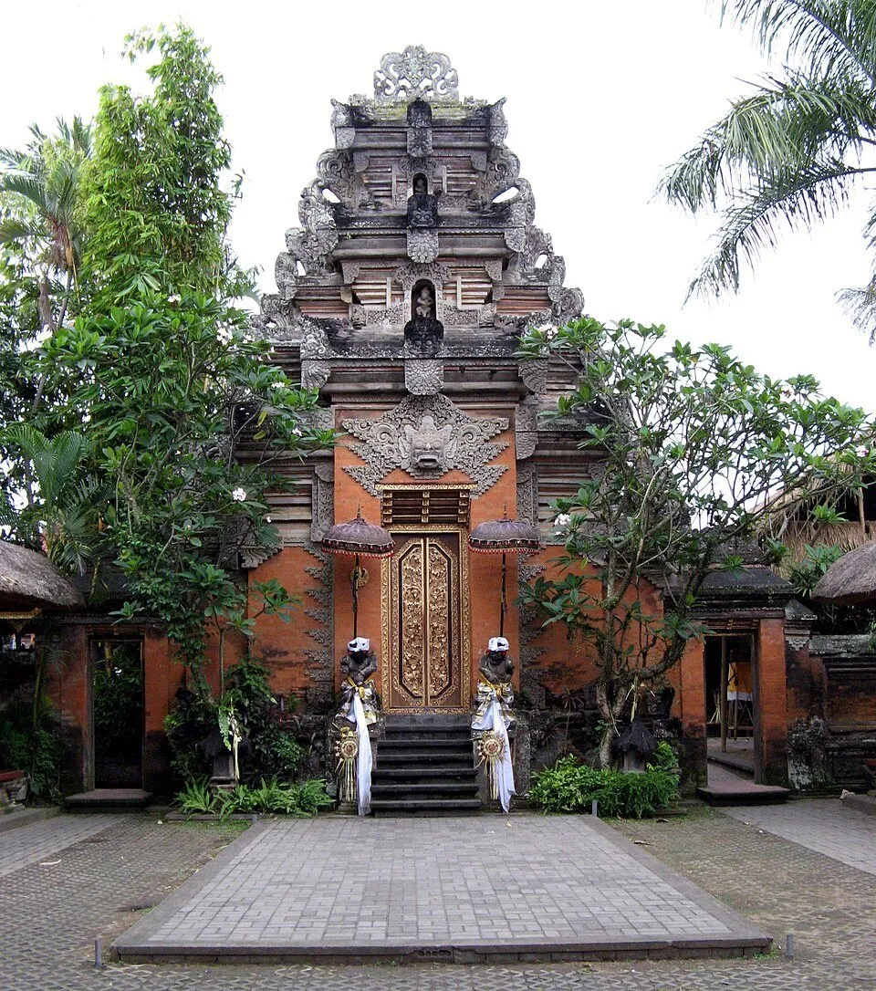 Portal esculpido do Palácio de Ubud com porta dourada, dois guardiões e degraus diante de um pátio.