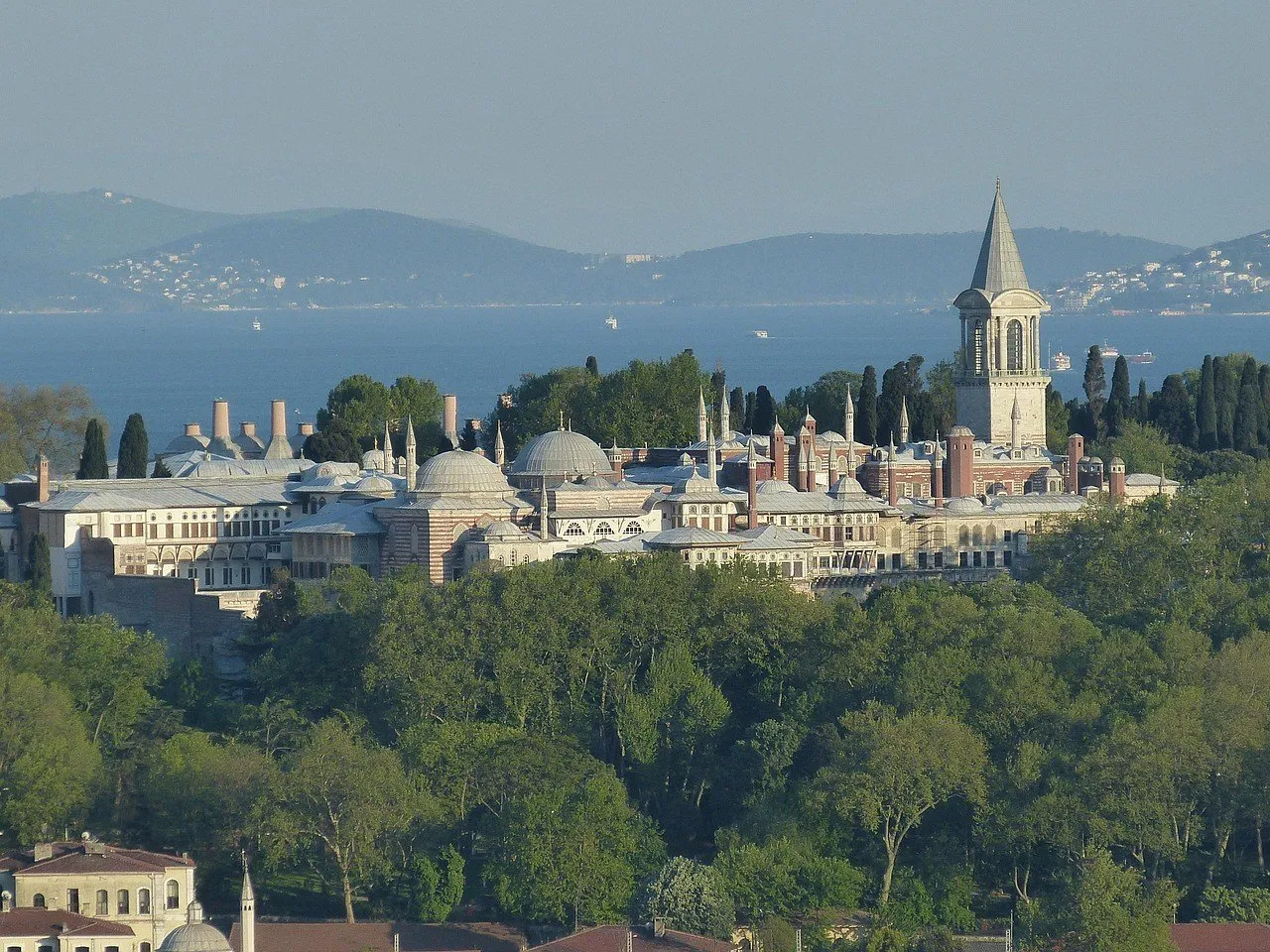 Conjunto do Palácio de Topkapı entre árvores, com a Torre da Justiça de teto cônico à direita e o mar ao fundo.