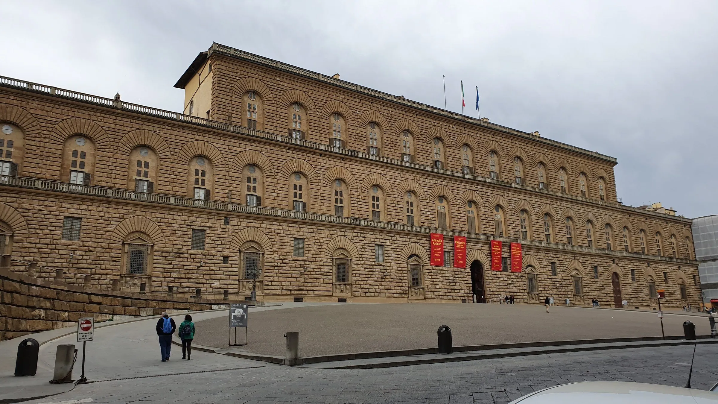 Fachada de pedra do Palazzo Pitti e praça inclinada, com poucas pessoas e céu nublado.
