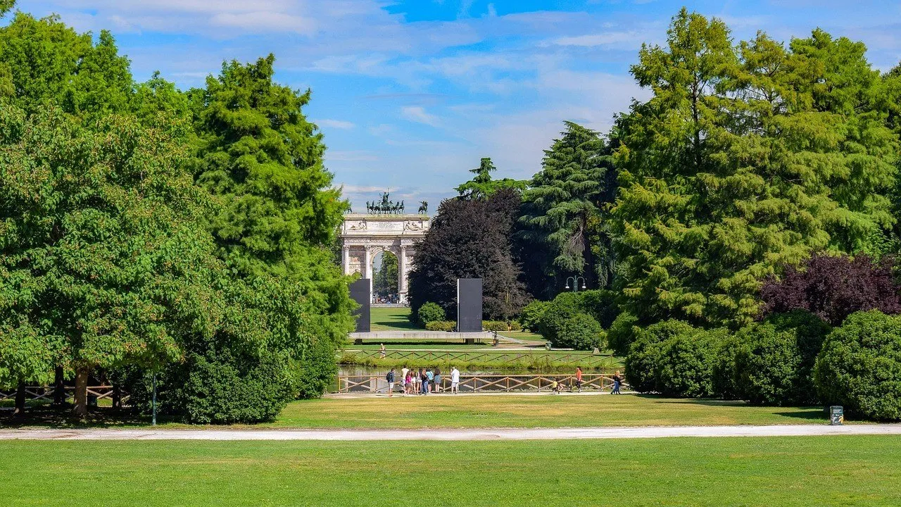 Gramados e árvores do Parco Sempione, com visitantes pequenos e o Arco della Pace ao fundo.