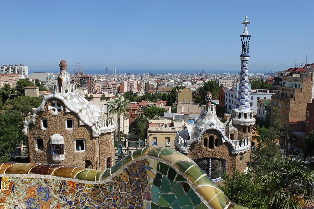 Banco de mosaicos do Park Güell em primeiro plano, pavilhões na entrada e cidade ao fundo.
