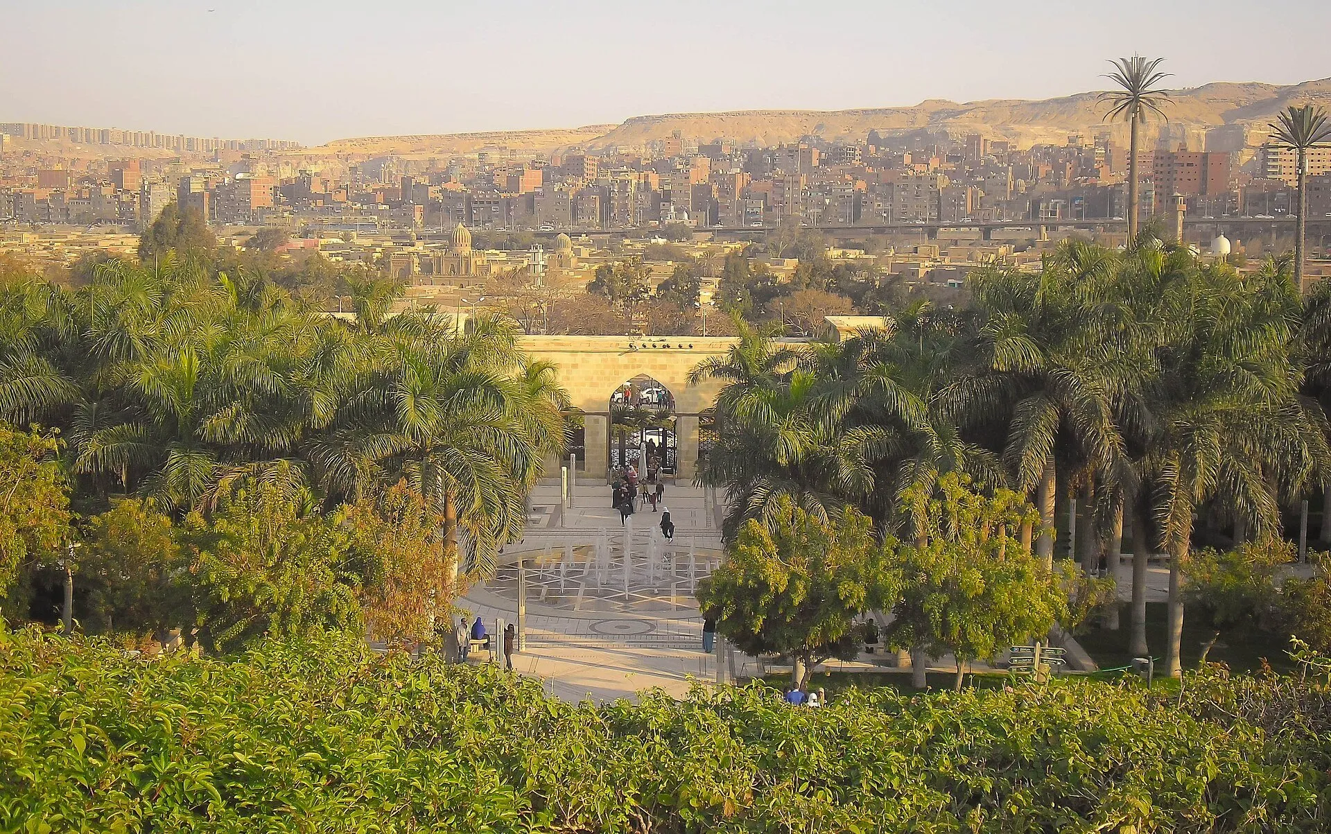 Caminho entre palmeiras no Parque Al-Azhar, com portal de pedra, visitantes e edifícios do Cairo ao fundo.