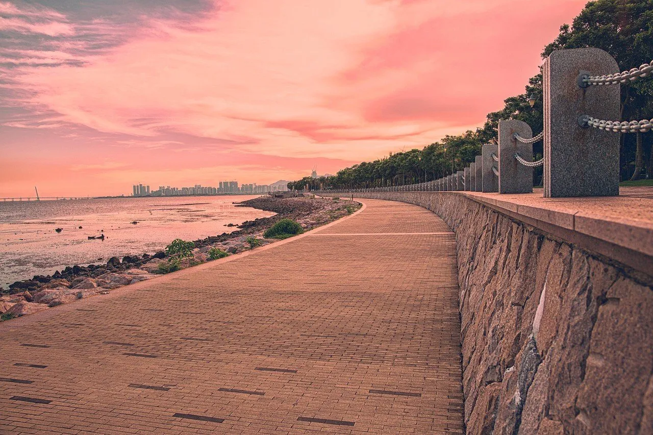 Passeio de tijolos curvo na orla da baía de Shenzhen, com muro de pedra e céu rosado do entardecer.