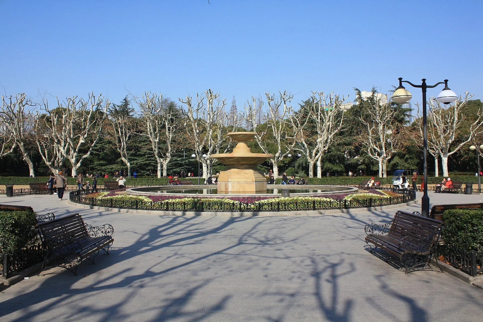 Fonte circular entre canteiros, bancos e árvores sem folhas no Parque Fuxing, sob céu azul.
