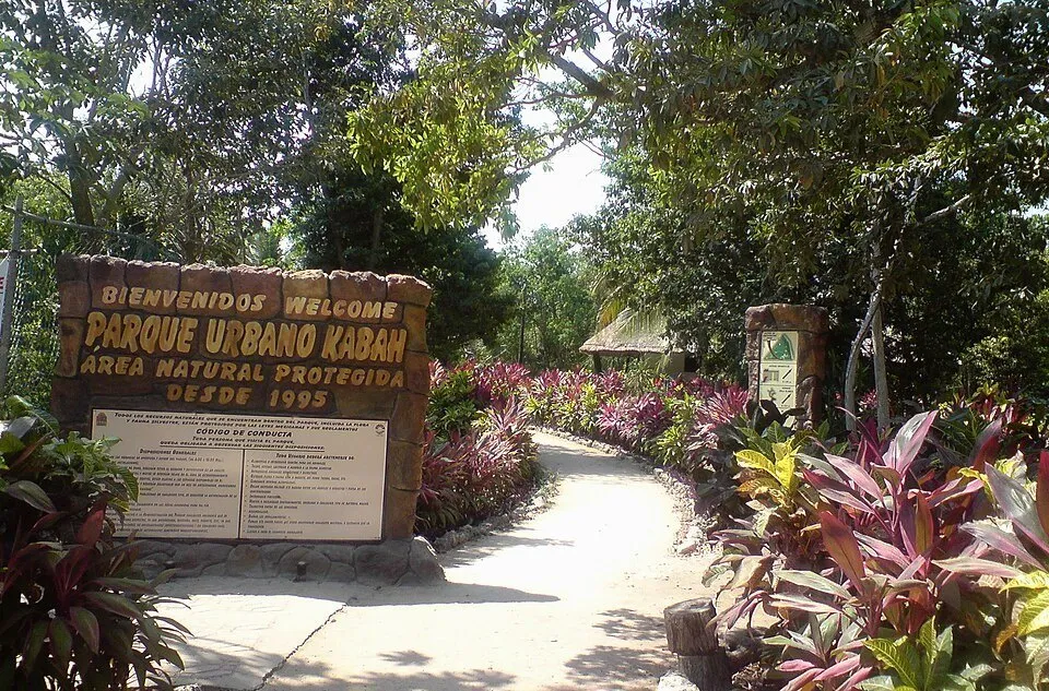 Entrada do Parque Urbano Kabah, com placa de madeira, caminho e vegetação colorida.