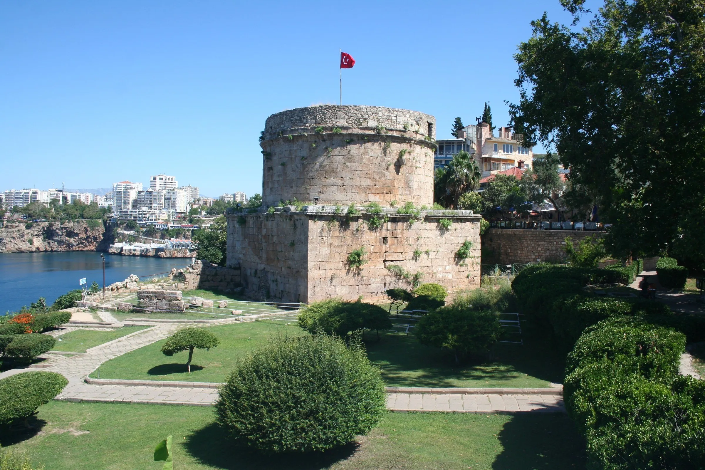 Torre Hıdırlık, de pedra, com base quadrada e corpo cilíndrico e uma bandeira turca, cercada por jardim com o mar ao lado.