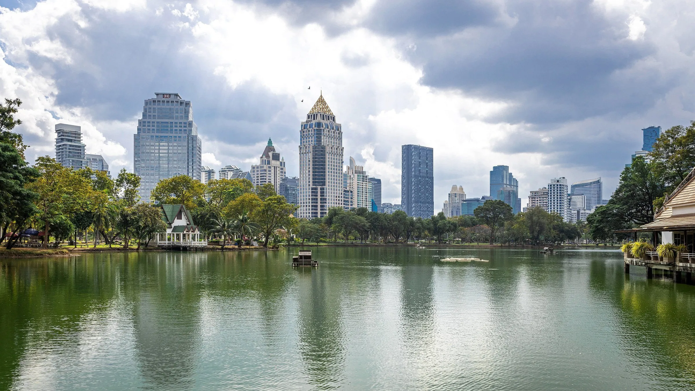Lago do Parque Lumphini, em Bangkok, com pavilhão à margem, árvores e arranha-céus ao fundo sob nuvens.