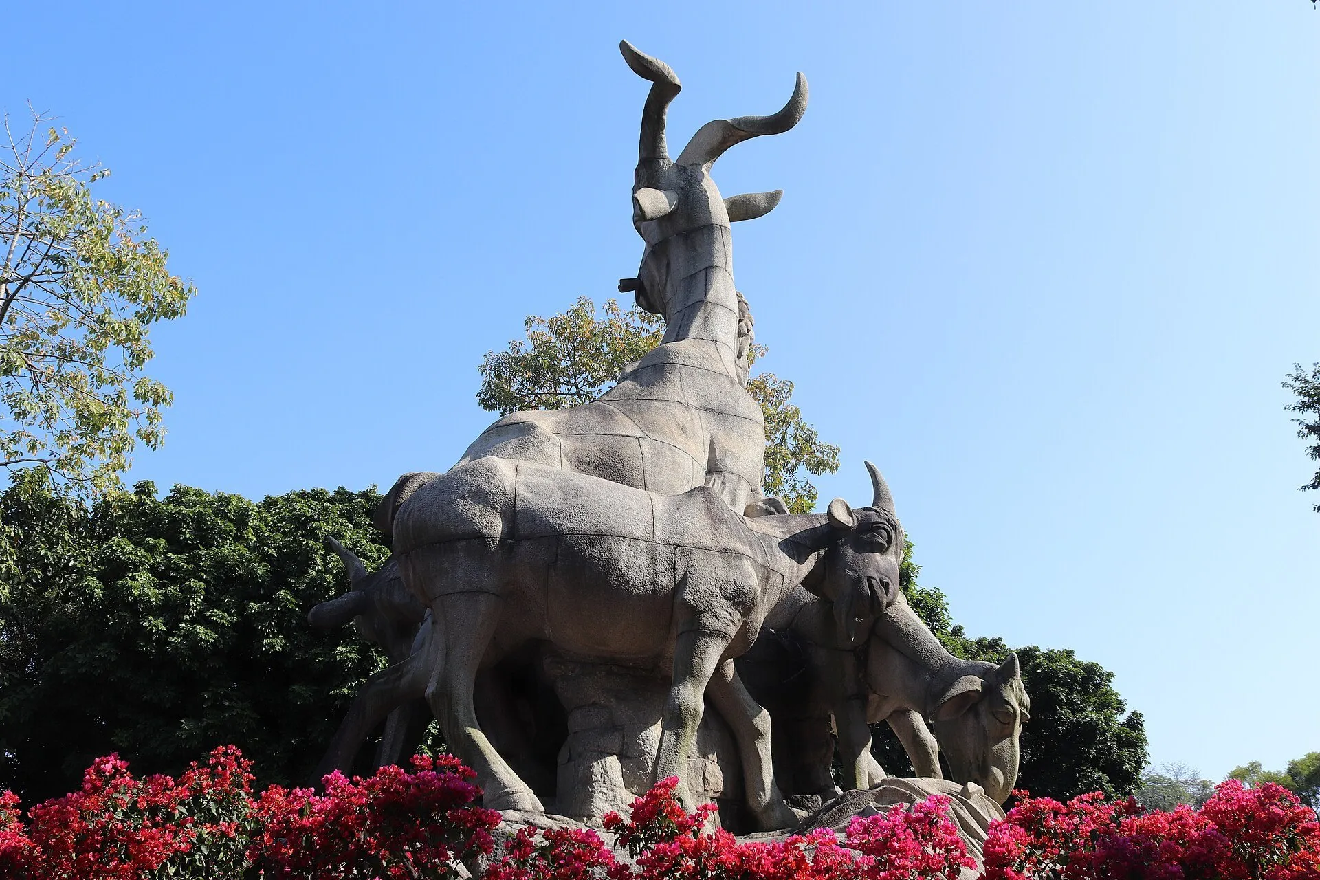 Escultura dos carneiros vista de lado no Parque Yuexiu, entre flores vermelhas, árvores e céu azul.