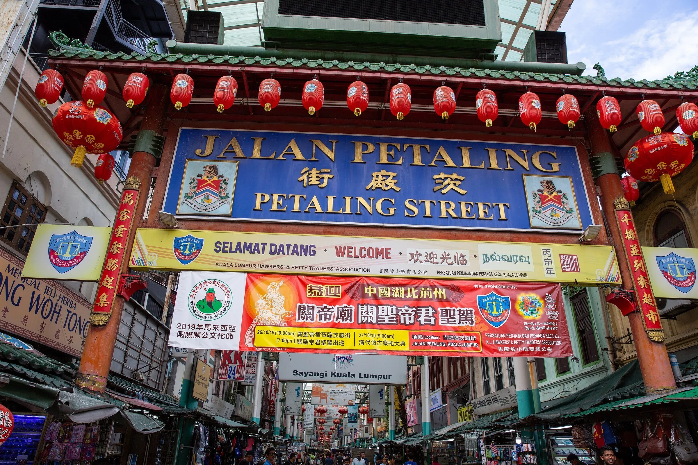 Portão chinês com letreiro de Jalan Petaling e lanternas vermelhas, com bancas e pessoas na rua coberta ao fundo.
