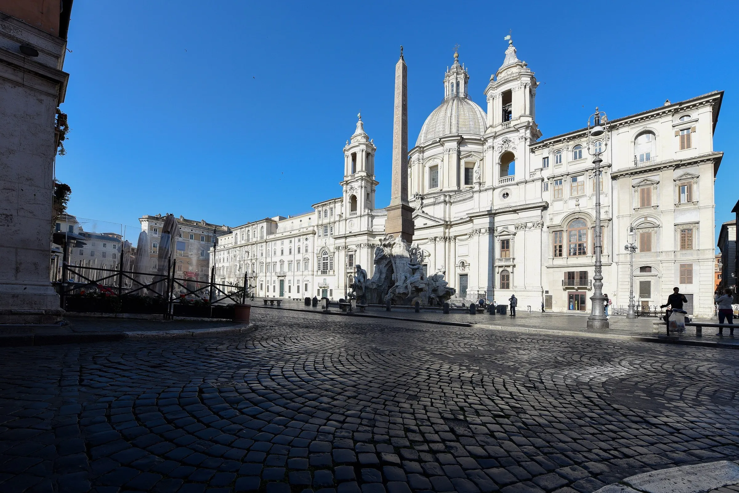 Praça de paralelepípedos com a igreja de Sant'Agnese in Agone, o obelisco e a Fonte dos Quatro Rios ao fundo, sob céu azul.