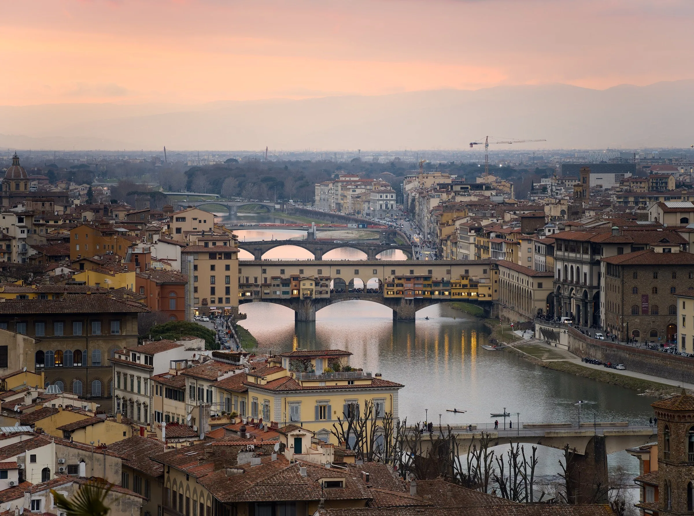 Vista elevada da Ponte Vecchio e de outras pontes sobre o Arno, com telhados e céu rosado.