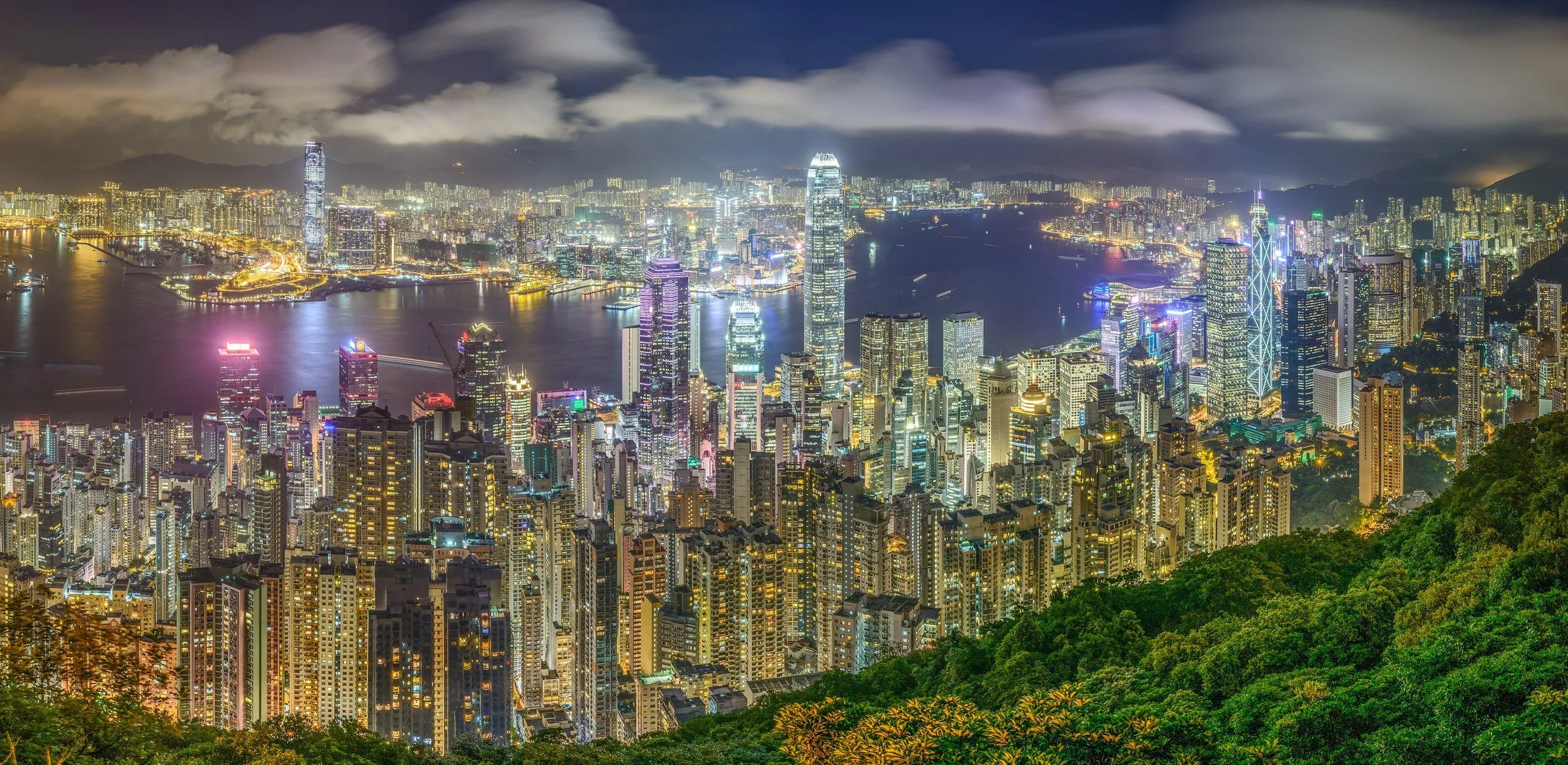 Vista noturna do alto do Pico Victoria sobre os arranha-céus de Hong Kong, o porto iluminado e a encosta coberta de mata.