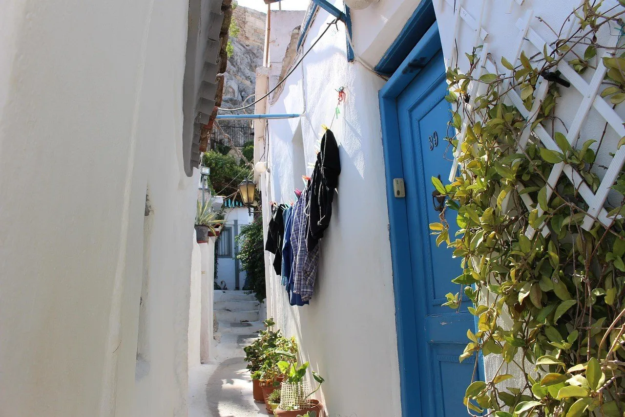 Passagem estreita em Plaka, Atenas, com paredes brancas, porta azul, plantas e degraus.