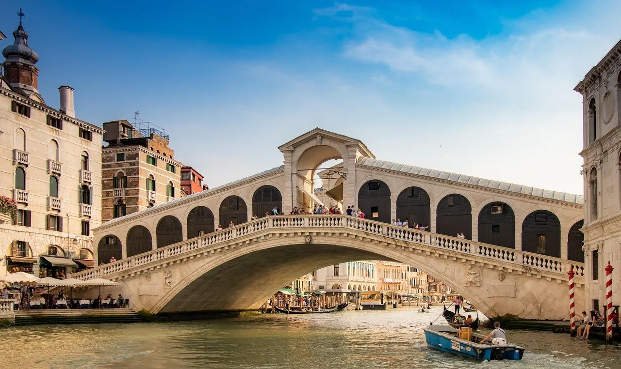 Arco de pedra da Ponte de Rialto sobre o Grande Canal, com visitantes e barcos passando sob a estrutura.