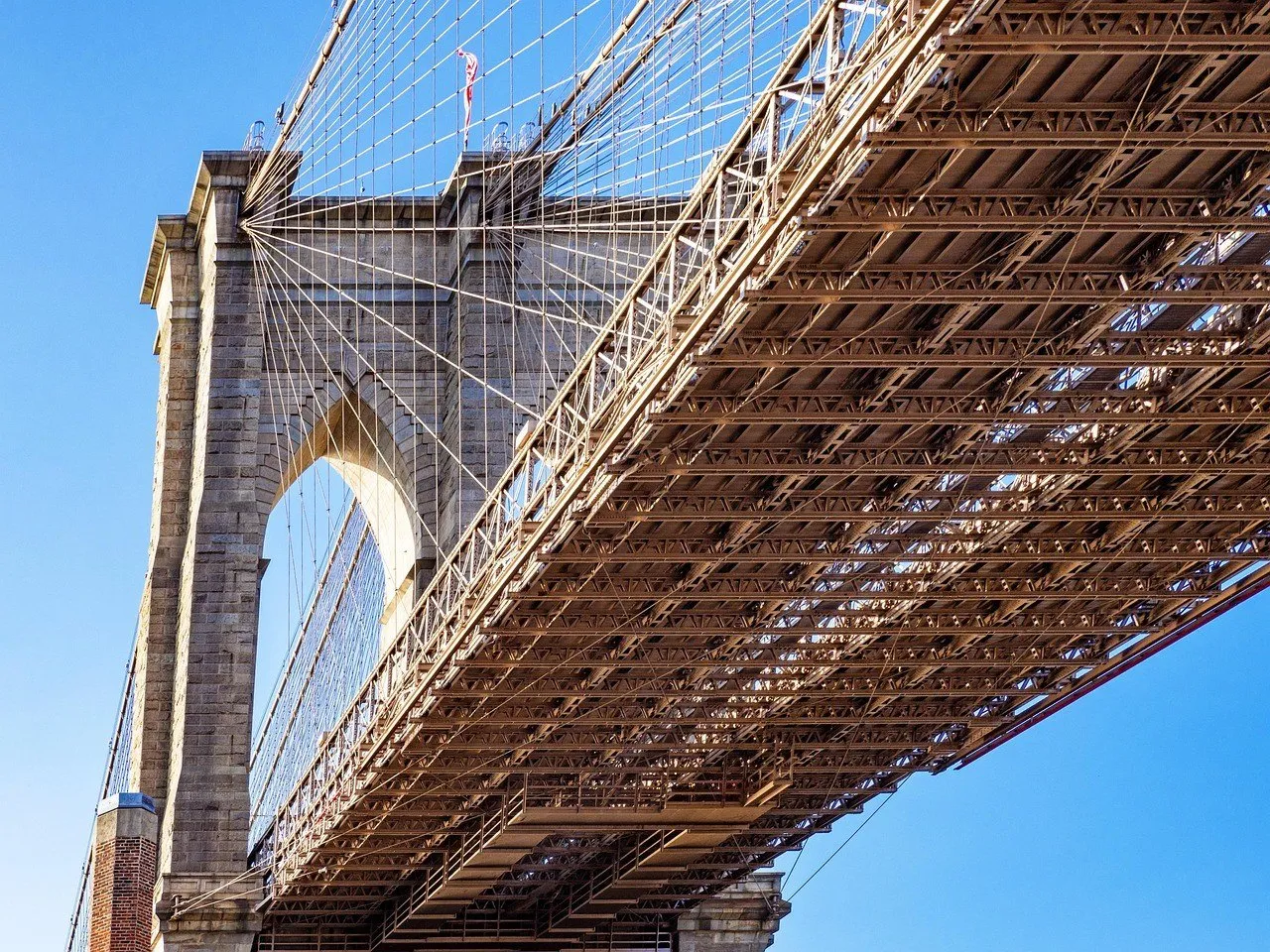 Torre de pedra com arcos góticos e o tabuleiro da Ponte do Brooklyn, vistos de baixo contra o céu azul.