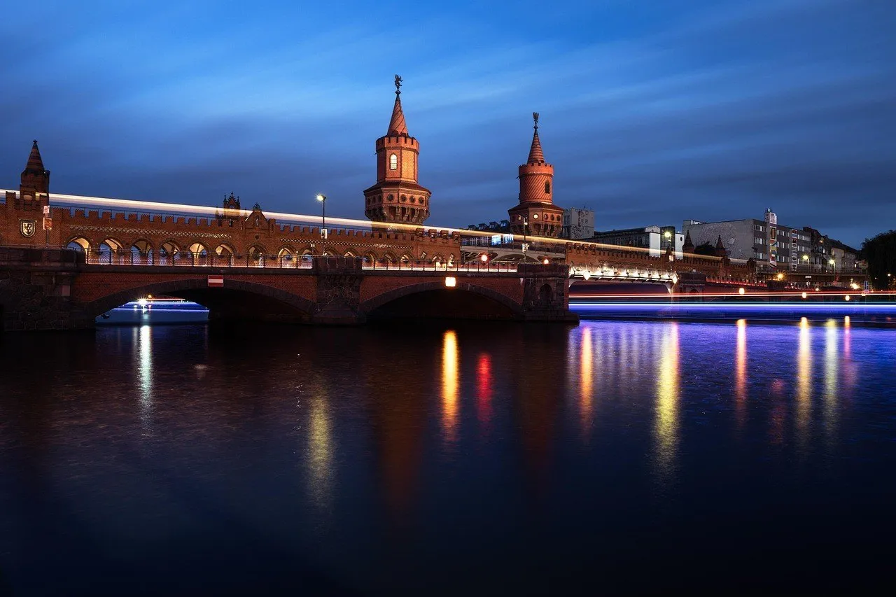 Ponte Oberbaum iluminada, com torres de tijolos, rastros de luz e reflexos no Spree.