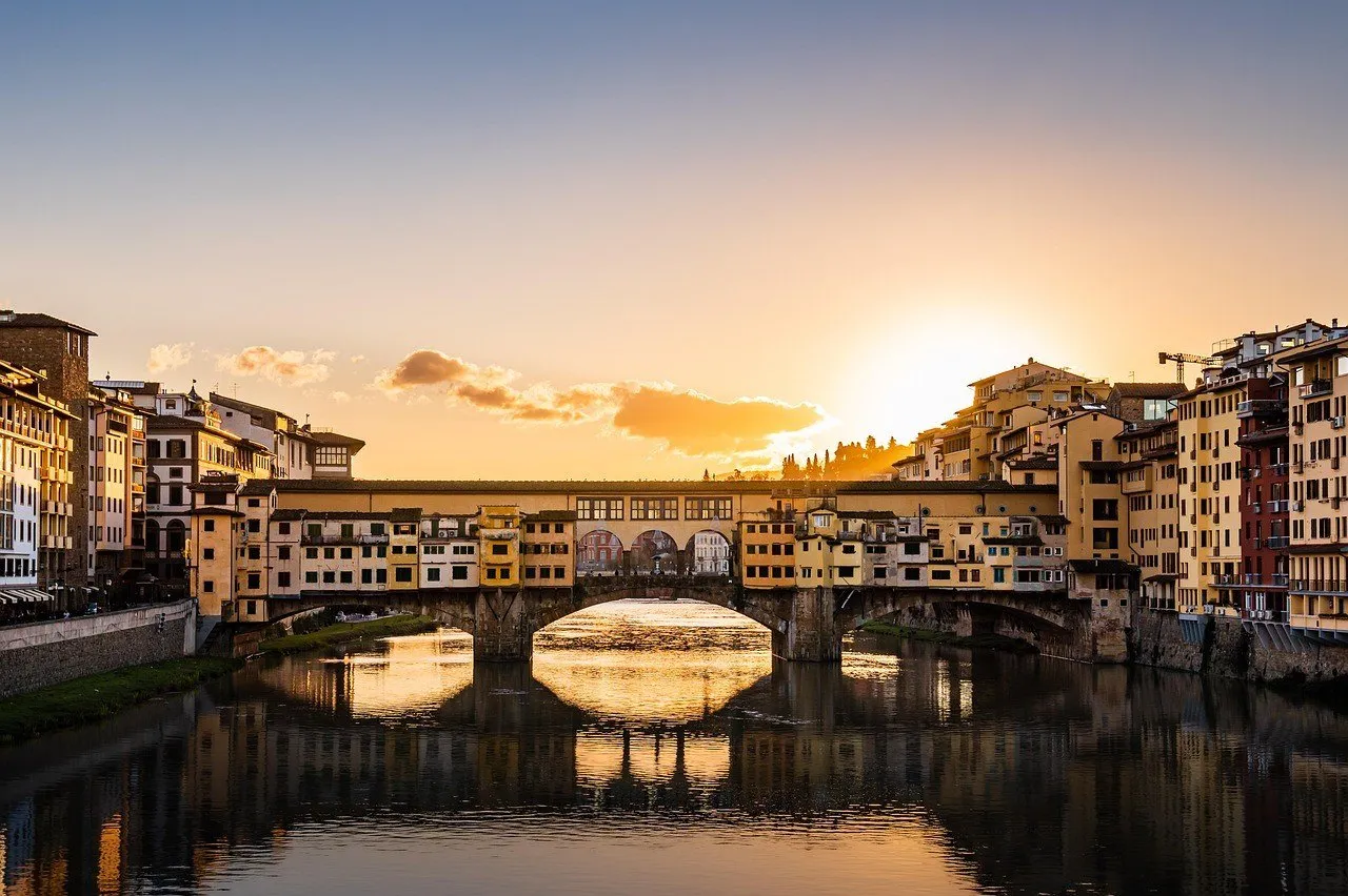 Ponte Vecchio vista de frente sobre o Arno, com reflexos dourados do pôr do sol entre os arcos.