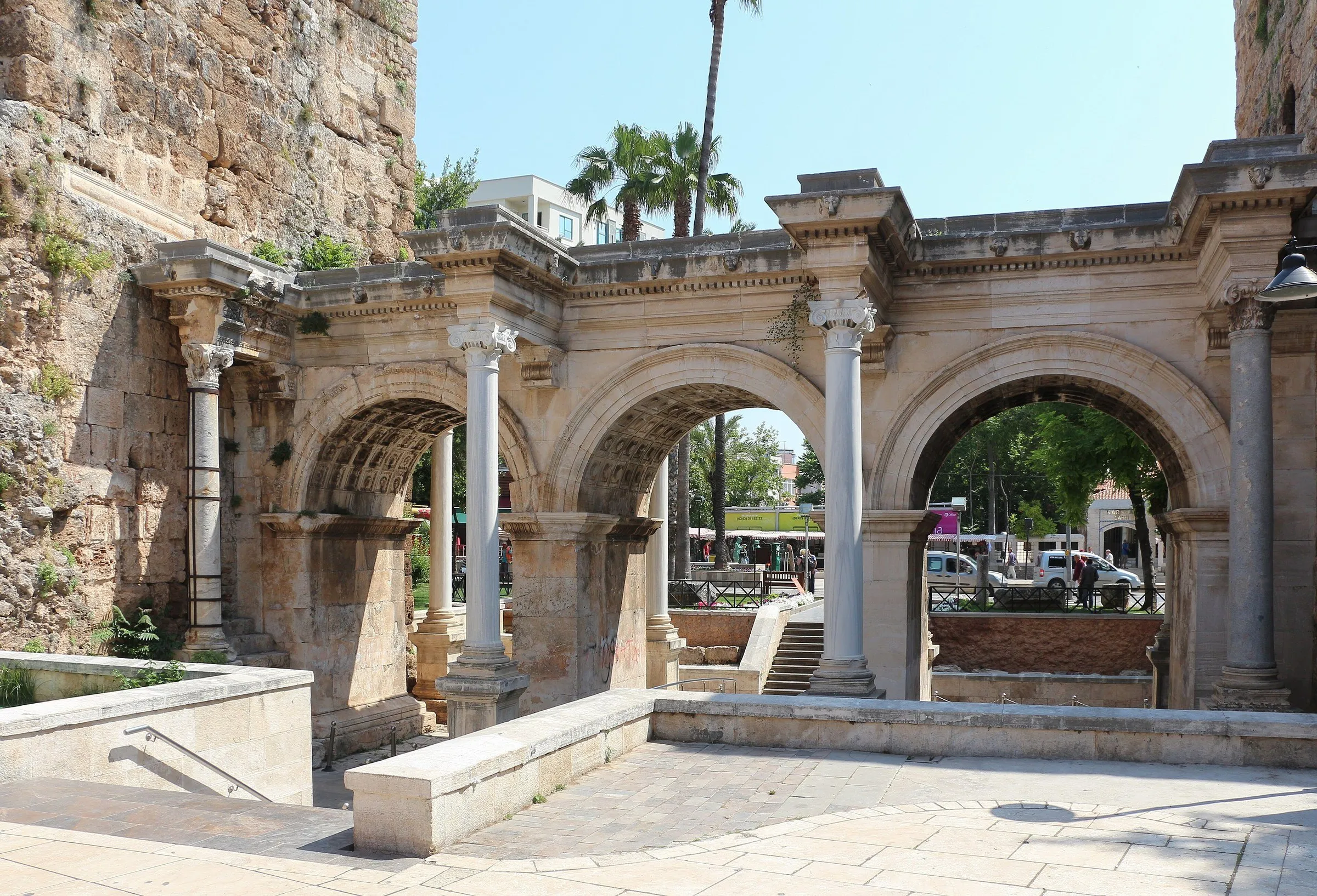 Portão de Adriano em Antalya, com três arcos de pedra e colunas de mármore, sob céu azul, ao lado de um trecho de muralha.