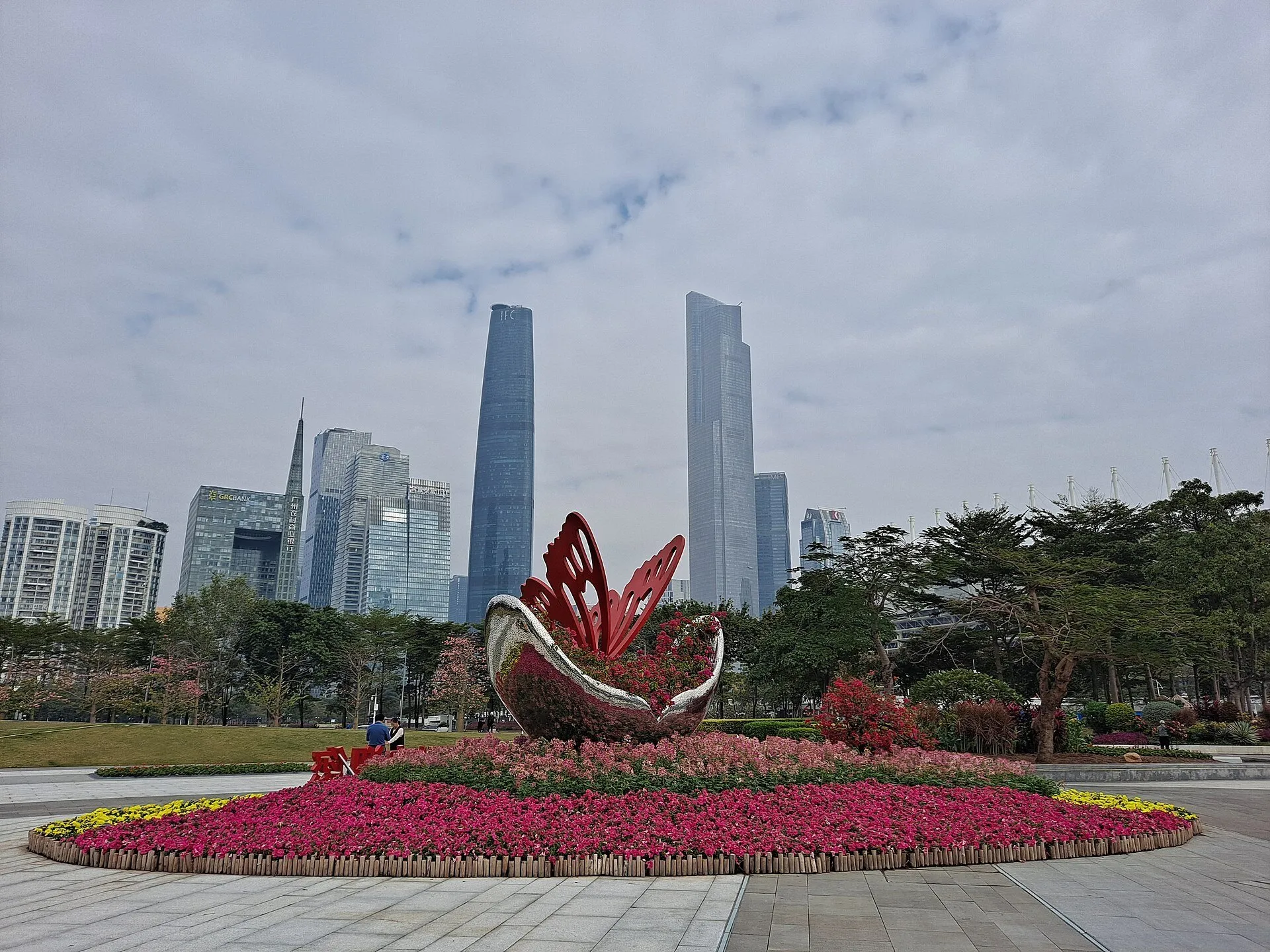 Canteiro de flores com escultura vermelha na Praça Huacheng, diante dos arranha-céus sob céu nublado.
