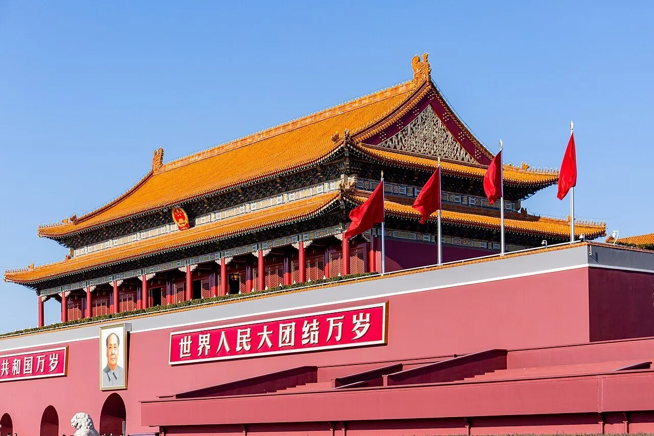 Portão Tiananmen em Pequim, com telhados amarelos, parede vermelha e bandeiras sob céu azul.