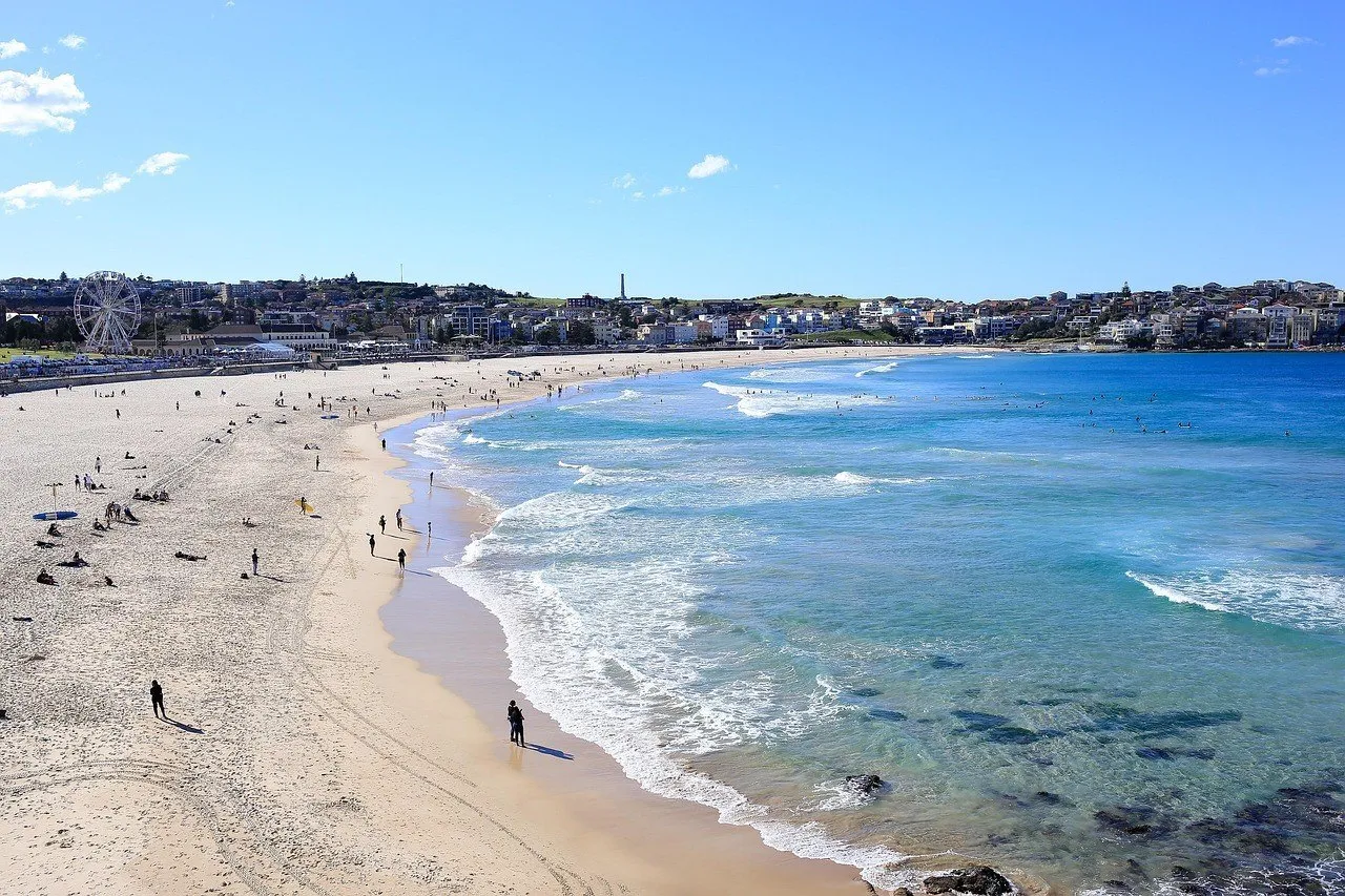 Curva da praia de Bondi, com areia clara, banhistas dispersos, ondas e prédios junto à orla.