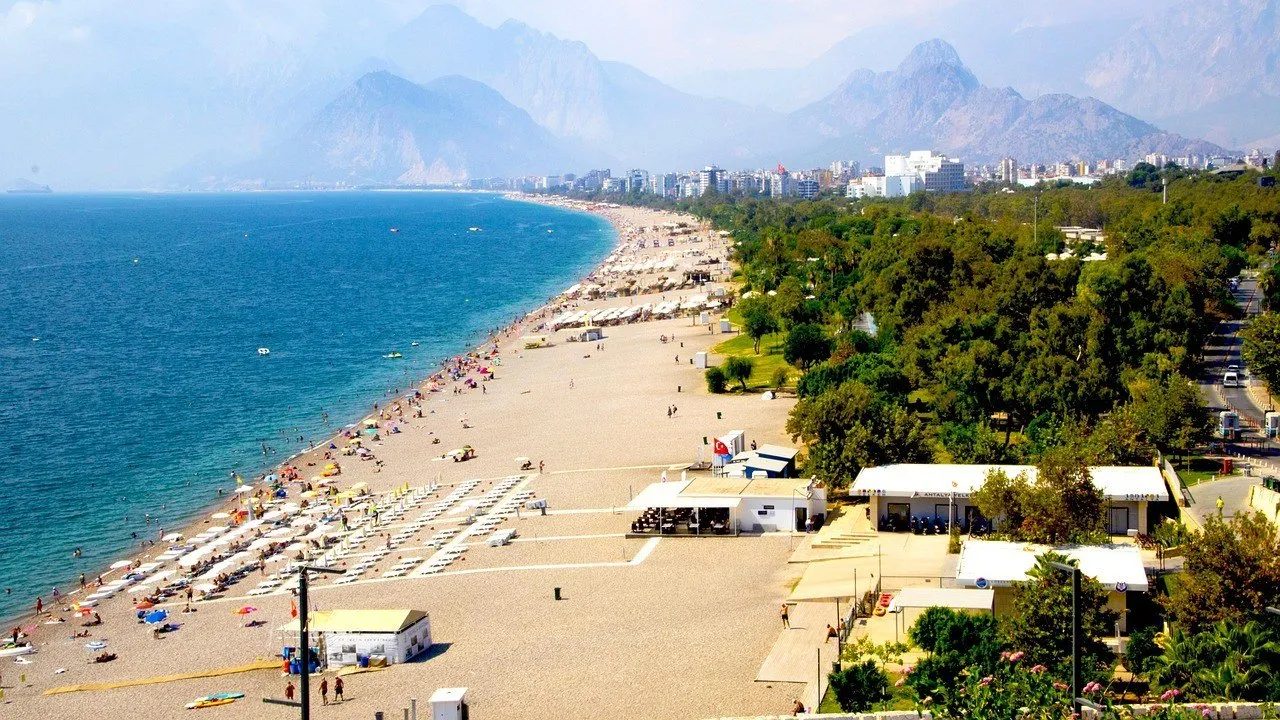 Vista aérea de Konyaaltı, com faixa de seixos, guarda-sóis, mar azul à esquerda, parque verde à direita e montanhas ao fundo.