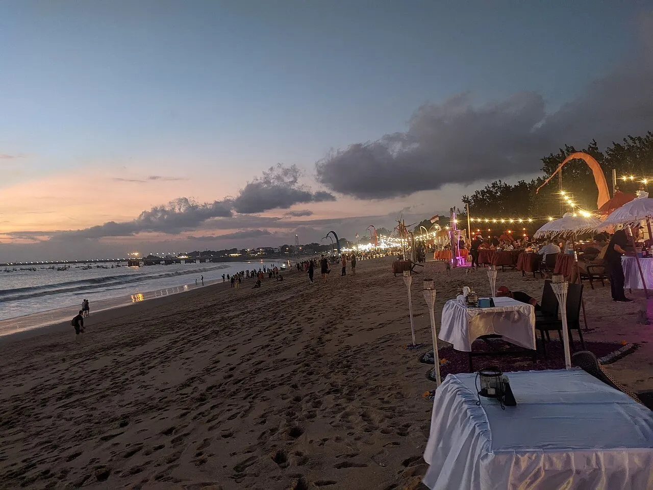 Mesas de restaurantes sobre a areia de Jimbaran ao entardecer, com cordões de luzes, mar e nuvens ao fundo.