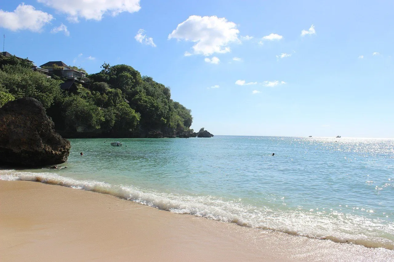 Areia clara e água azul-esverdeada da praia de Padang Padang, com rochas e vegetação na margem esquerda.