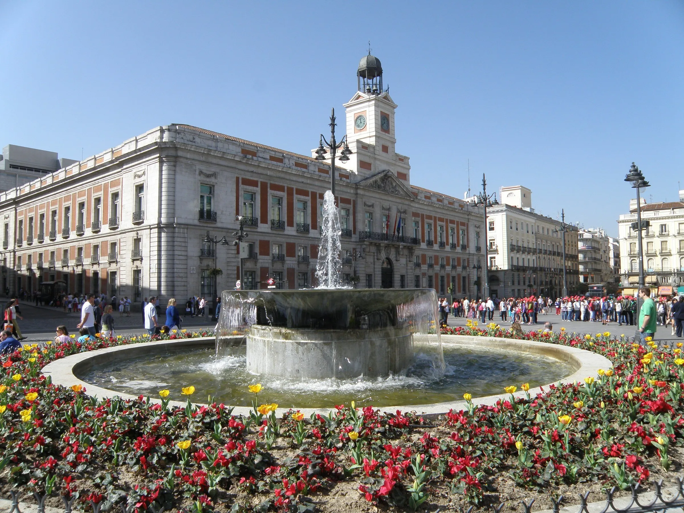 Casa de Correos e seu relógio na Puerta del Sol, com fonte, flores e pedestres à frente.
