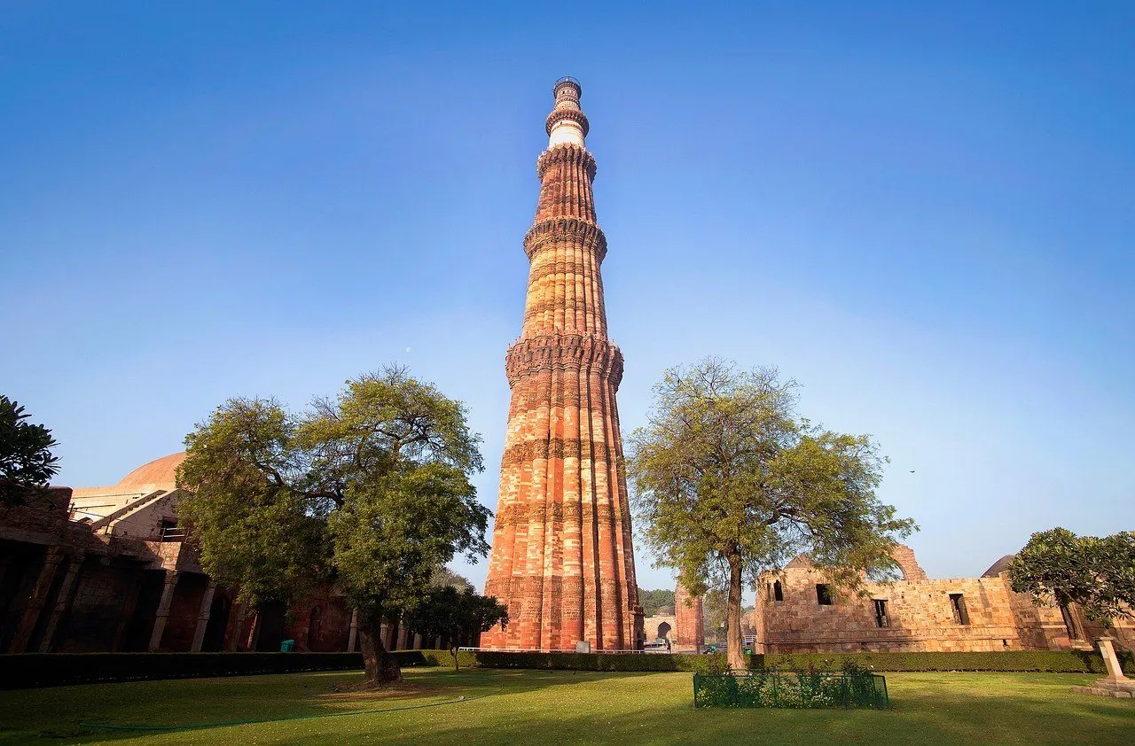 Torre de arenito vermelho do Qutub Minar, em Délhi, com faixas de inscrições, entre árvores e ruínas, sob céu azul limpo.